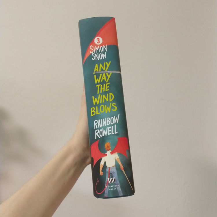 Any Way the Wind Blows by Rainbow Rowell - book spine shows the title and under that, Simon facing away from us with a red dragon tail and wings, holding a sword