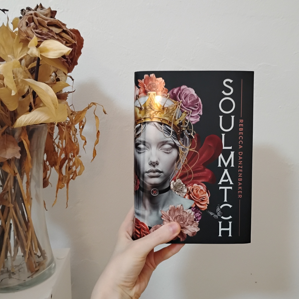 Soulmatch by Rebecca Danzenbaker hardcover. The cover is what looks like a marble carving of a young woman's face and neck, wearing a gold crown, with red and pink flowers all around and behind face. Background is black.