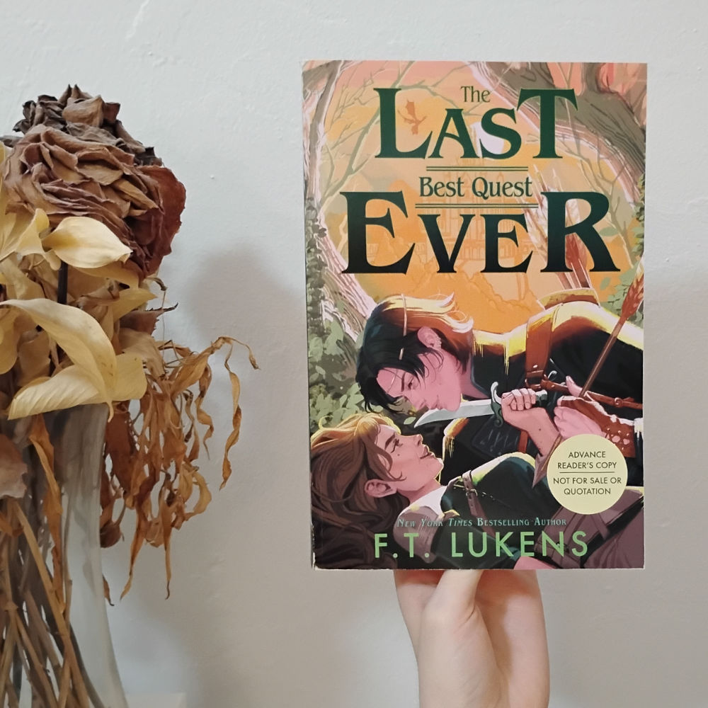 Physical ARC of The Last Best Quest Ever by FT Lukens. Cover is artwork of an androgynous black-haired person in royal clothes somewhat on top of a brown-haired girl in adventuring clothes. The royal is holding an arrow to the girl's chest, and the girl is holding a knife to the royal's throat, both looking more flirty than threatening.