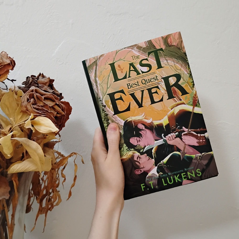 Photo of the hardcover of The Last Best Quest Ever by FT Lukens. Cover is artwork of an androgynous black-haired person in royal clothes and ear piercings somewhat on top of a brown-haired girl in adventuring clothes. The royal is holding an arrow to the girl's chest, and the girl is holding a knife to the royal's throat, both looking more flirty than threatening.