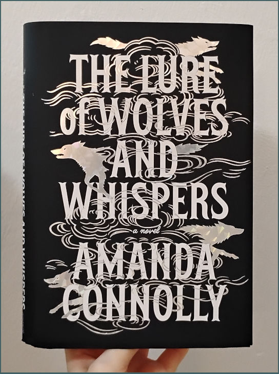Photo of the hardcover of The Lure of Wolves and Whispers by Amanda Connolly. The title and author take up the whole cover, capitalized in a clear but stylized text. Lines like wind swirl through and around the text, and partial silhouettes of wolves are coming out of the wind on the sides of the text. The background is black, the text and images white/silver.